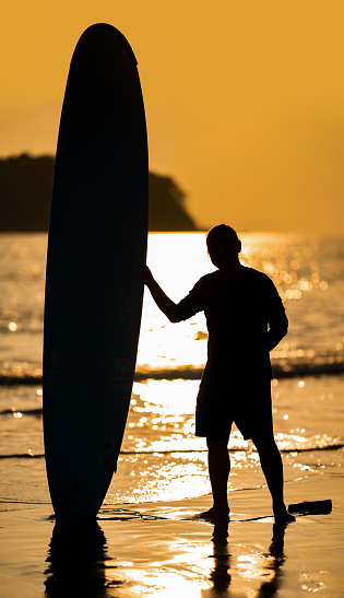 Silhouette Smart Asian Man long hair touches and poses with Standed Surfboard on the beach in Twilight time