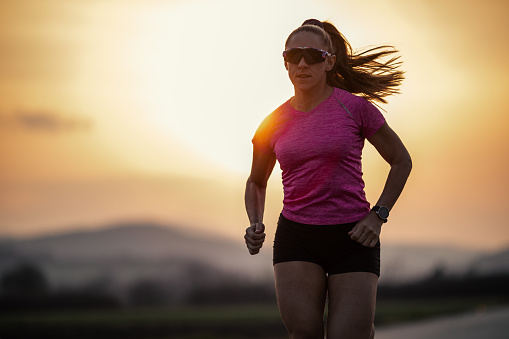 Mid adult woman jogging over empty road at sunset. She is marathon runner