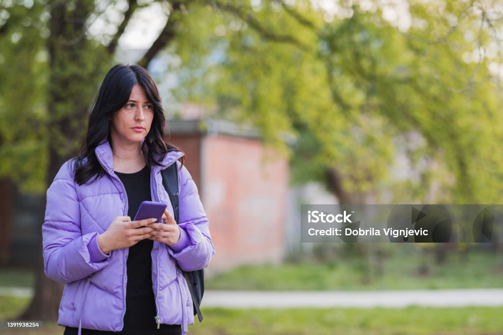 Girl holding smart phone when standing in the park Young woman in purple winter jacket standing in the park and holding smart phone Acne Stock Photo