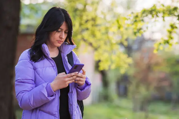 Photo of Young woman in purple jacket standing in park and using phone