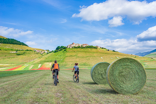 Couple cycling mtb in blooming cultivated fields of Castelluccio di Norcia highlands, famous colourful flowering plain in the Apennines, Italy. Agriculture of entil crops and red poppies.