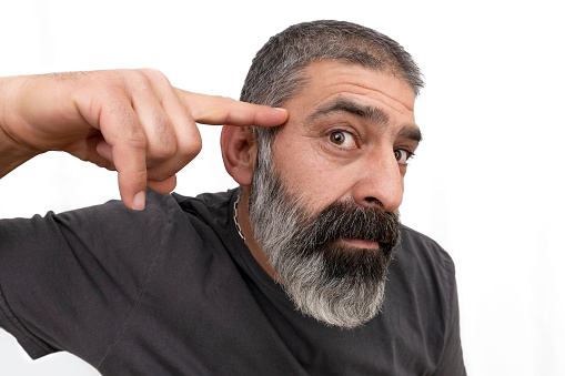 Mature beared man looking at camera against textured white background.\nHe points with his finger at his brain to explain how to use his brain.