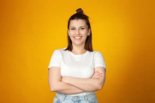 Studio portrait of an attractive 24 year old woman with long brown hair in a white t-shirt and blue jeans on a yellow background