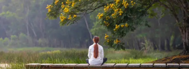 Photo of Panorama back view of woman relaxingly practicing meditation in the public park to attain happiness from inner peace wisdom with yellow flower blossom in summer