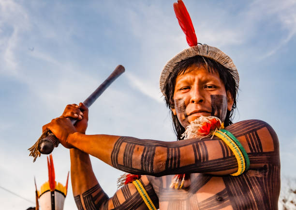 Indigenous man with body tribal paint Indigenous man from an Amazon tribe in Brazil with tribal art painted on his body and face, wearing a headdress made of straw and a red feather and holding a stick. 2009. indigenous peoples day stock pictures, royalty-free photos & images