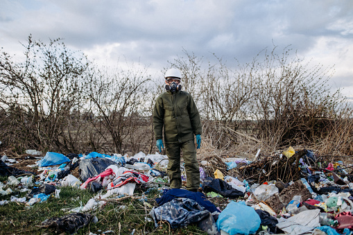 The woman in a protective suit and respirator standing among plastic pollution in nature