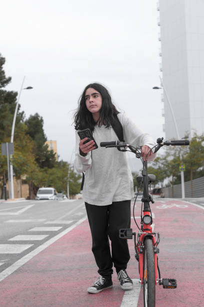 Vertical image of a teenager walking along a bike path pushing a bike while consulting his smart phone. Vertical image, low angle, long shot, of a teenager walking along a bike path pushing a red bicycle while consulting his smart phone. He has long hair and looks to the side. trans boy stock pictures, royalty-free photos & images