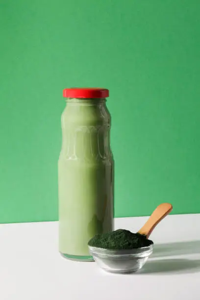 Photo of green smoothie in bottle and spirulina algae powder in glass bowl on green, white background.useful habits,self care and healthy lifestyle concept.