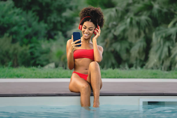 joven afroamericana con el pelo rizado escuchando música y mirando la pantalla del teléfono inteligente junto a la piscina. joven en traje de baño y grandes auriculares rojos que se relajan al aire libre junto a la piscina - tourist resort audio fotografías e imágenes de stock