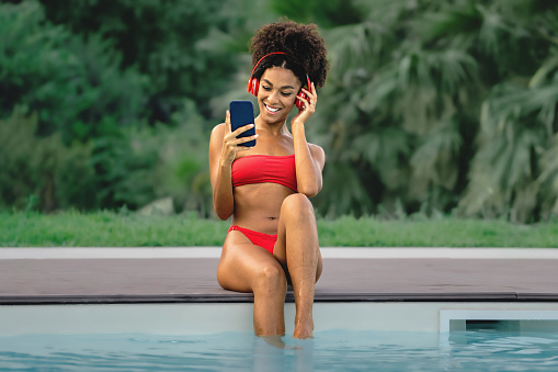 African American young woman with curly hair listening music and looking at smartphone screen by the pool. Young lady in swimsuit and big red earphones chilling outdoors poolside