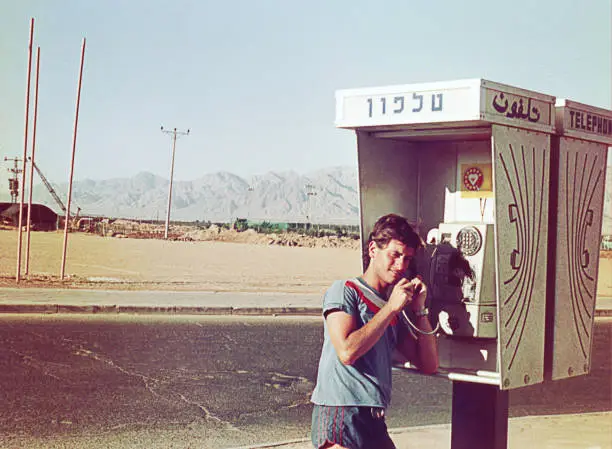 Grainy analog photo of a young man talking by phone in a public phone booth in the middle of nowhere. Analog image from the eighties.