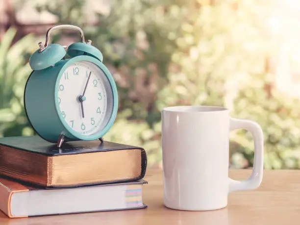 Photo of Alarm clock on workbook and textbook on the table or work desk with hot coffee in home office. Workplace and lifestyle business.