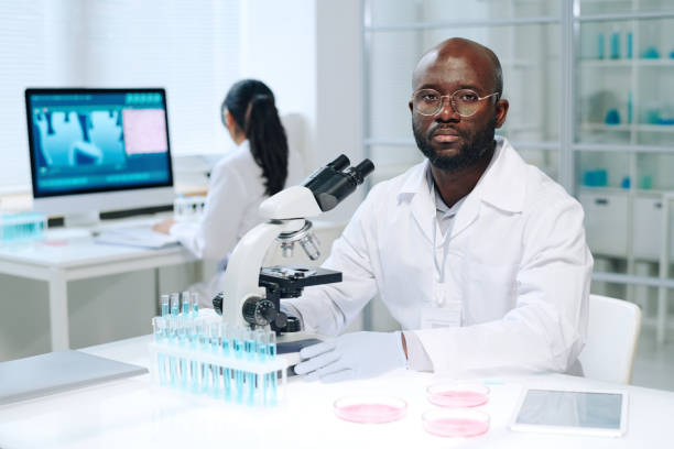 young serious scientist in lab coat sitting by workplace in front of microscope - scientist research test tube lab coat imagens e fotografias de stock