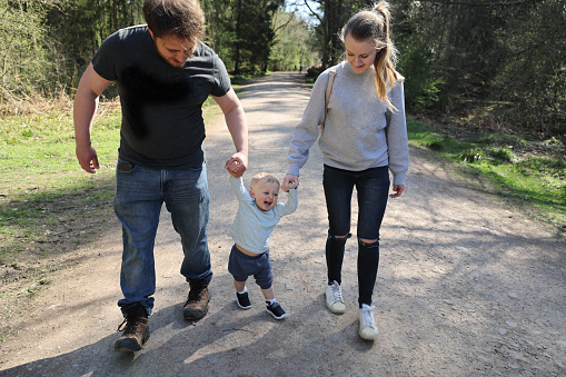 Young Mum and Dad swinging their happy, laughing 18 month old daughter between them on a sunny forest walk