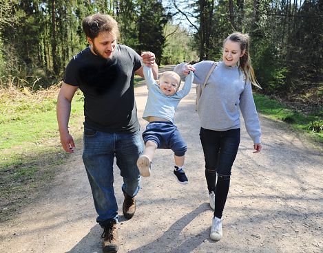 Young Mum and Dad swinging their happy, laughing 18 month old daughter between them on a sunny forest walk