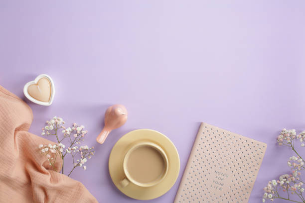 Cute girls workspace with morning coffee cup, pink notebook, beige scarf, heart shaped candle, gypsophila flowers on pastel purple background. Love, romance concept.