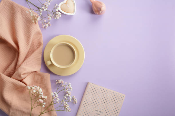 Morning coffee, heart shaped candle, pink notebook, beige scarf, decor and spring flowers on pastel purple table. Romantic breakfast still life. Flat lay, top view.