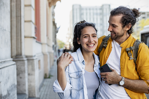 Cheerful heterosexual couple walking and laughing. Young male is embracing his girlfriend and holding mobile phone. Beautiful female is looking at the camera.