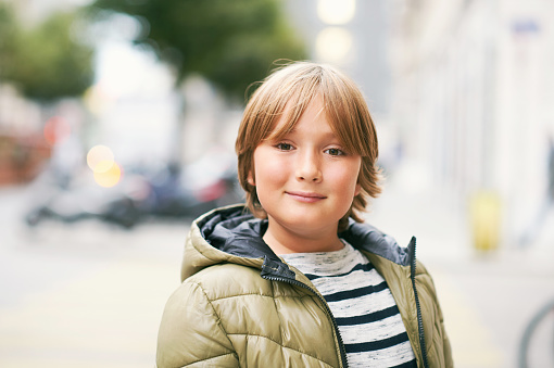 Outdoor portrait of handsome 10 year old kid boy, wearing warm jacket, looking straight at camera, posing on city street