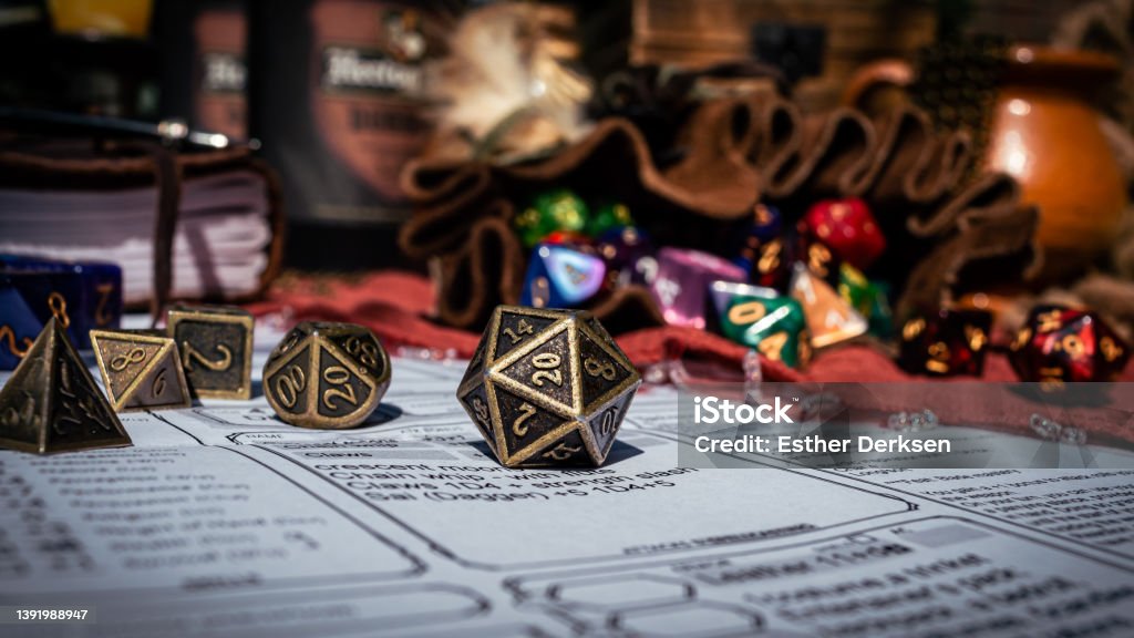 Low angle image of a brass d20 Low angle image of a brass 20-sided die on a character sheet in the sun. In the background are a dice bag and a notebook Board Game Stock Photo