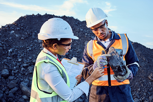 Two young African mine workers wearing protective wear are discussing coal quality while on site at a coal mine