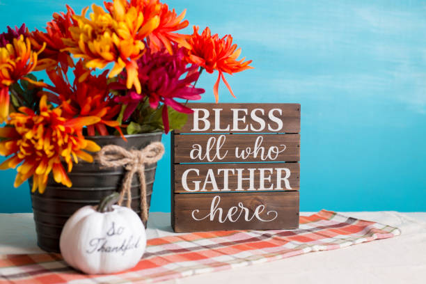 Autumn table decorations.  Bucket of orange flowers,  turquoise background and plaid table runner or cloth.  White pumpkin.  Bless all who Gather here on wooden box. stock photo