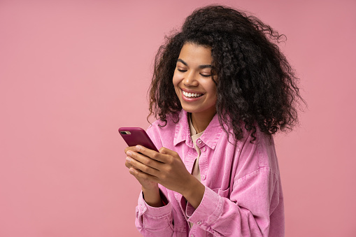 Beautiful smiling African American woman using smartphone playing mobile game isolated on pink background. Happy stylish female holding mobile phone shopping online with sale, copy space
