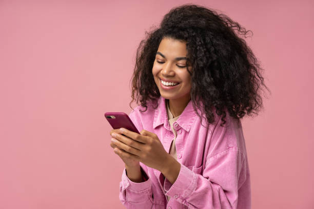 Smiling African American woman using smartphone playing mobile game isolated on pink background. Happy stylish female holding mobile phone shopping online with sale stock photo