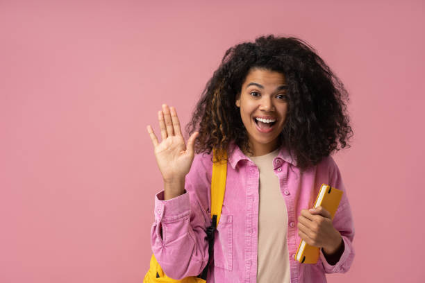 Excited African American student with backpack and book greeting, looking at camera isolated on pink background. Back to school, education concept stock photo
