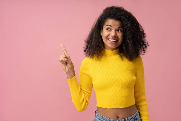 Young smiling African American girl wearing stylish yellow sweater pointing finger on copy space isolated on pink background. Shopping, sale, advertisement concept stock photo