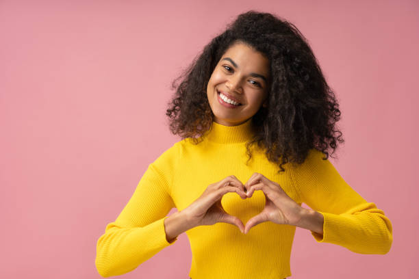 Smiling African American woman with toothy smile showing heart shape near face isolated on pink background stock photo