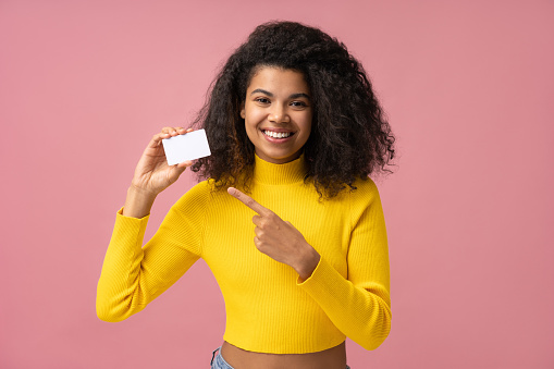 Portrait of beautiful smiling curly haired woman holding blank business card pointing finger on copy space isolated on pink background. Mockup, advertisement concept