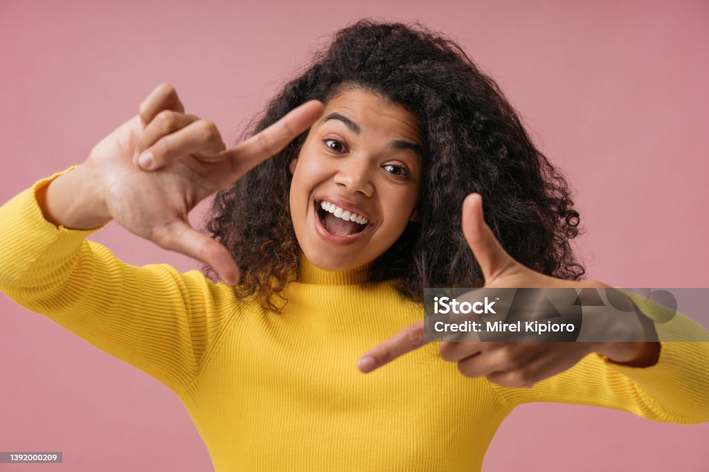 Emotional African American woman with toothy smile holding hands near face looking at camera. Excited photographer making frame isolated on pink background Beautiful emotional African American woman with toothy smile holding hands near face looking at camera. Excited photographer making frame isolated on pink background Mouth Open Stock Photo