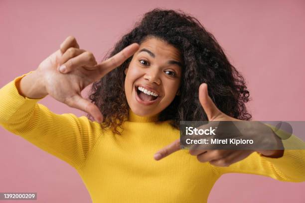 Emotional African American Woman With Toothy Smile Holding Hands Near Face Looking At Camera Excited Photographer Making Frame Isolated On Pink Background Stock Photo - Download Image Now