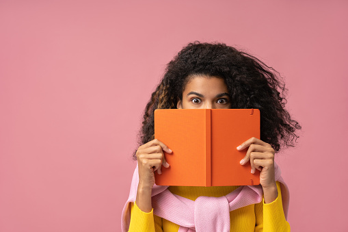 Young pensive African American student holding book near face isolated on pink background, copy space. Education concept