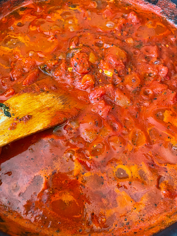 Stock photo showing elevated view of a rich tomato sauce steaming on a cooker hob, in a large frying pan being stirred with a wooden spatula as it reduces.