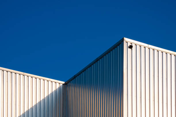 青く澄んだ空の背景に対する工業ビルの段ボール鋼壁の表面の太陽光反射 - corrugated iron ストックフォトと画像