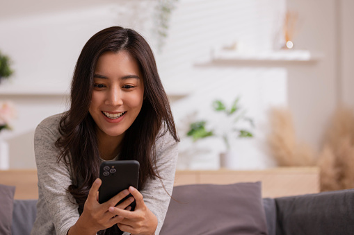 At home, an Asian woman waving a smartphone app enjoys online virtual video chat with pals in a virtual meeting while sharing stories for social media.