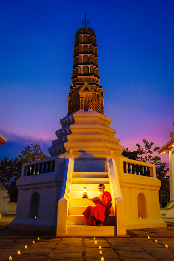 Thai Buddhist monk or novice sitting at buddhist pagoda in buddhism temple reading and studying Buddhist lessen book at night