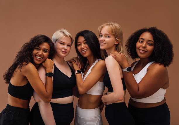 Five women of different body types embracing together. Cheerful females in sport clothes posing on brown background. stock photo