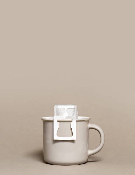 istock Drip coffee bag with ground coffee in big cup, package with paper bag drip coffee filter for brewing on beige color background, side view with copy space, monochrome color 1392069576