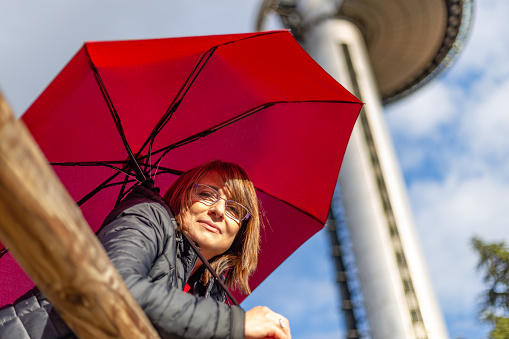 Madrid, Spain. March 7 2022. Low angle view of middle-aged woman with red parasol and glasses looking at camera in sunny afternoon. In the background, communications tower. Insurance, bank accounts, future, pensions, retirement