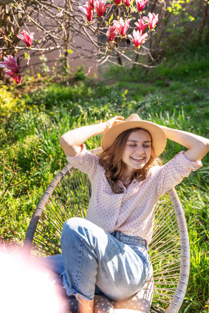 咲き誇る庭園に座りながら春を楽しむ帽子をかぶった若く美しい幸せそうな笑顔の女性 - sunlight flower magnolia flower head ストックフォトと画像
