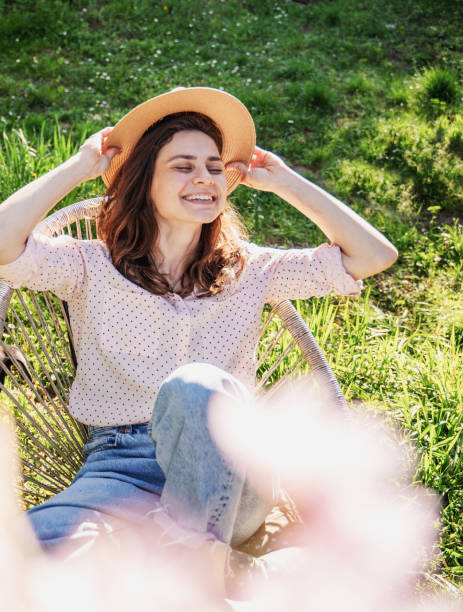 joven hermosa mujer sonriente feliz con un sombrero disfrutando de la primavera mientras está sentada en un jardín en flor - plant white magnolia tulip tree fotografías e imágenes de stock