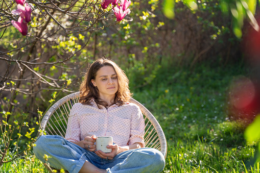 Young beautiful woman enjoying relaxing in the spring while sitting in the blooming garden of her home with a cup of tea