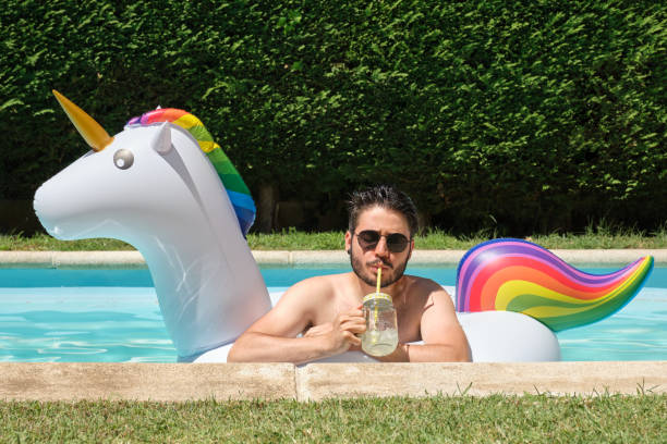 istock Young man with sunglasses drinking lemonade into a unicorn inflatable ring. 1392075881