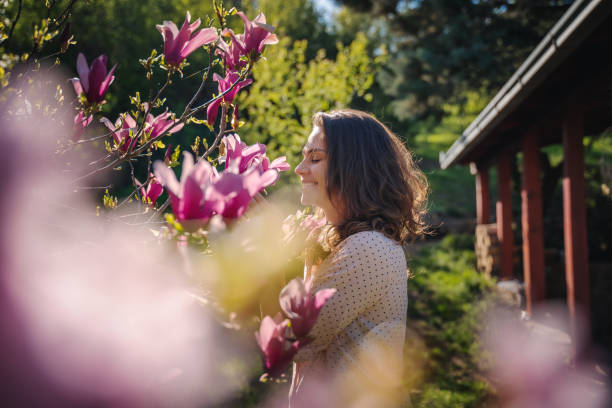 花の香りを吸い込む咲き誇るモクレンの木の近くの庭で自宅の春を楽しむ若い美しい女性 - sunlight flower magnolia flower head ストックフォトと画像