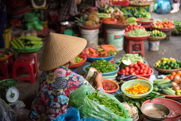 Vietnamese street vendor with traditional conical hat (Non La) selling vegetables Hoi An, Vietnam - May 07, 2018: Woman selling vegetables at Hoi An Central Market (Cho Hoi An) in the morning. vietnam stock pictures, royalty-free photos & images