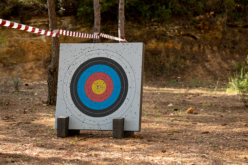 Archery range in the woods. Shooting target full of holes from arrow shots. Arrow on the ground and archery target with different scoring zones highlighted in black, blue red and yellow in the center.