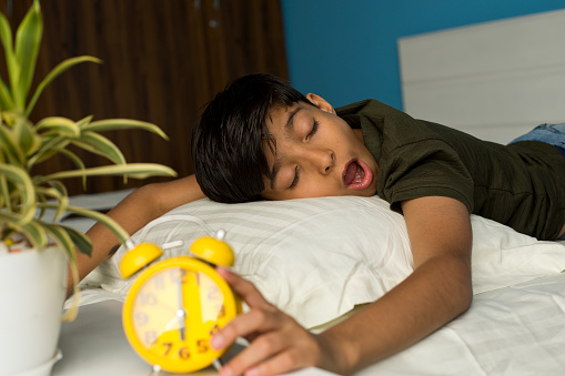Boy reaching over to turn off alarm clock while sleeping on bed